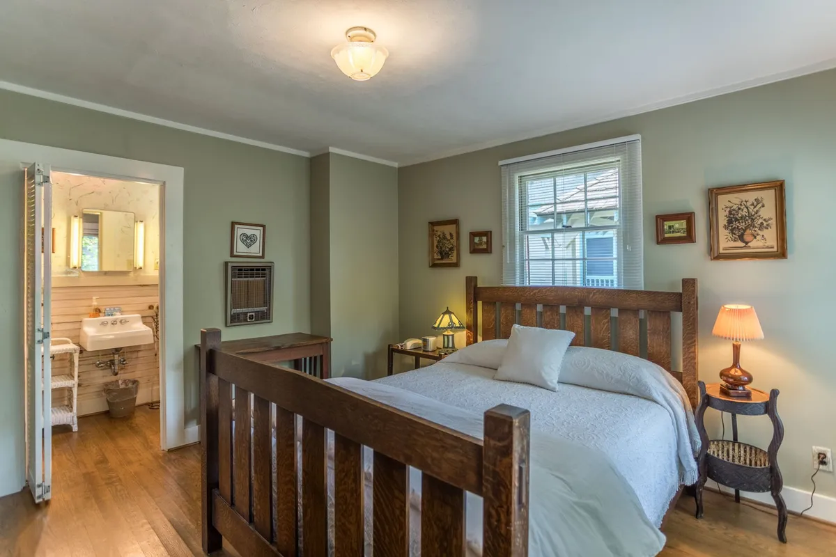 A restful bedroom featuring a comfortable bed dressed in soft linens, a nightstand with a warm lamp, subtle artwork above the headboard, and an open doorway that leads into an ensuite bathroom. Sunlight streams through the window, casting a serene glow across the wooden furnishings and the cozy setting.