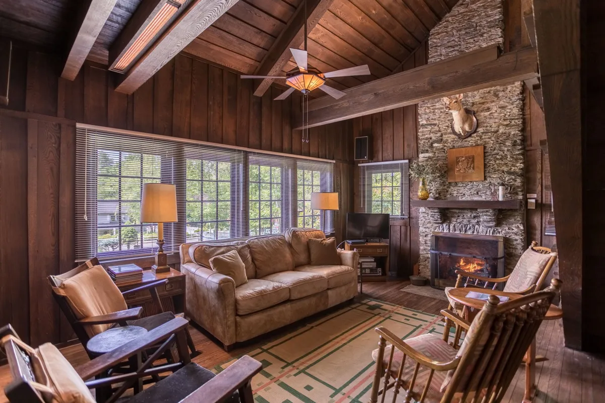 This image captures a sun-drenched living room centered on a striking floor-to-ceiling stone fireplace. The fireplace mantel displays decorative items, while large windows fill the space with natural light. A comfortable sofa and visible wooden beams enhance the warmth and authenticity, evoking the inviting atmosphere of a Mission-style bungalow.