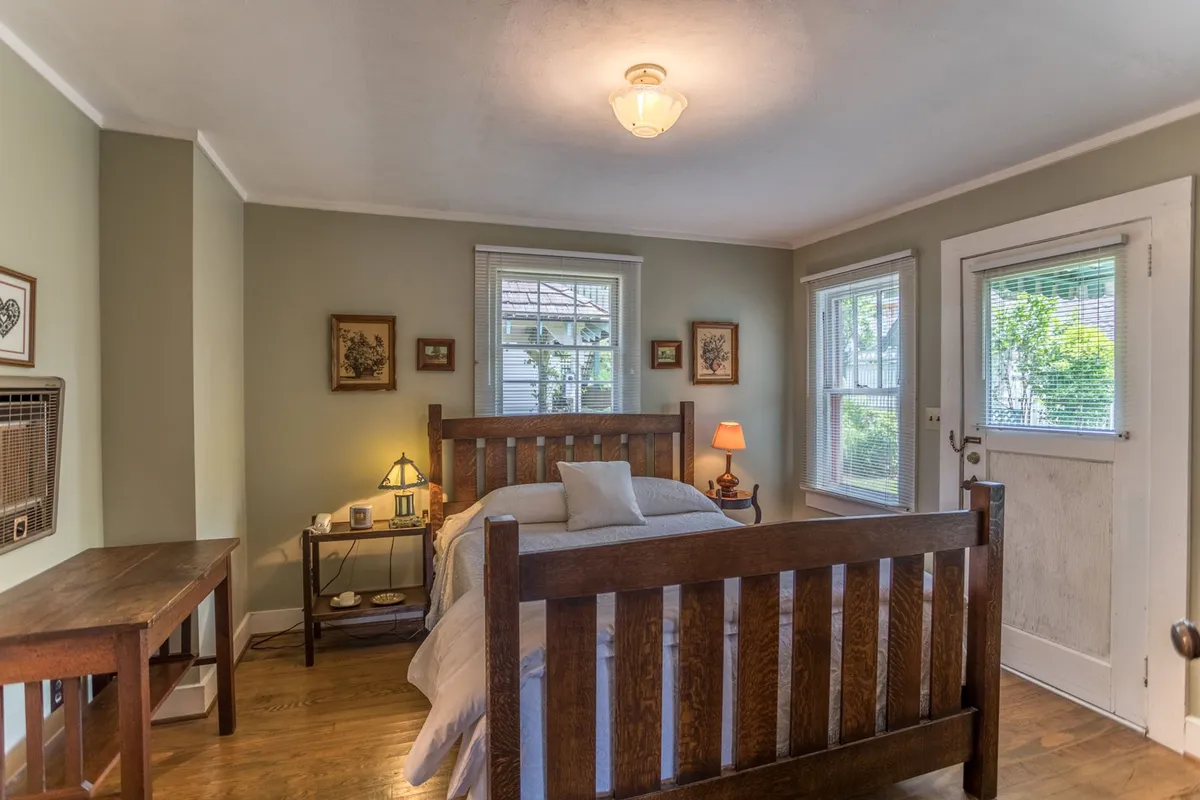 A warmly lit bedroom featuring a custom wooden bed topped with cozy linens. A small bedside lamp and artwork on the walls create a welcoming ambiance, while a window offers a glimpse of greenery outside, hinting at the peaceful garden retreat beyond.