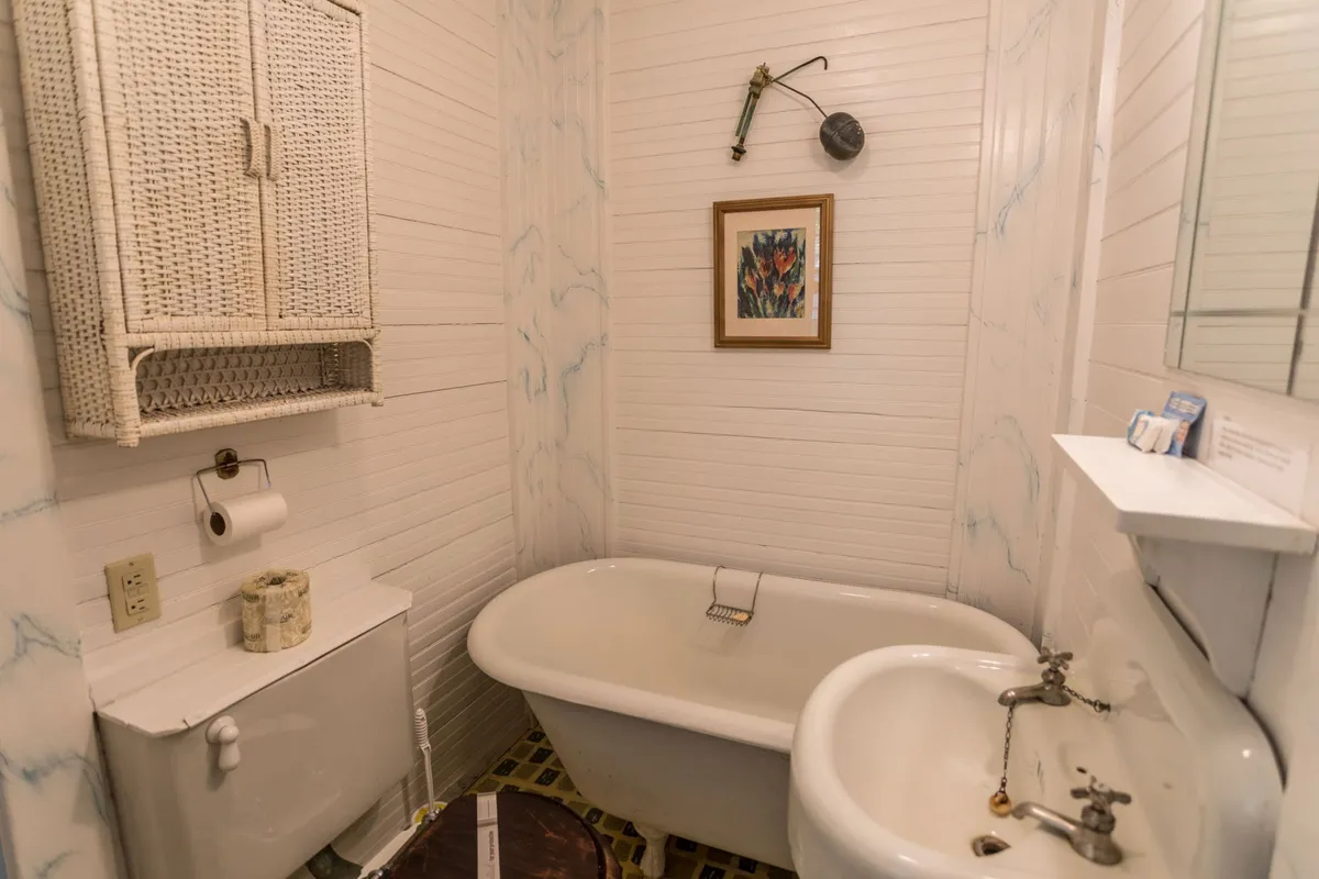 A tranquil bathroom scene featuring a claw-foot bathtub, vintage sink, woven cabinet, and period-appropriate wall art. The lighting is soft, inviting restful moments and self-care routines, perfectly aligned with the home's lovingly restored historic character.