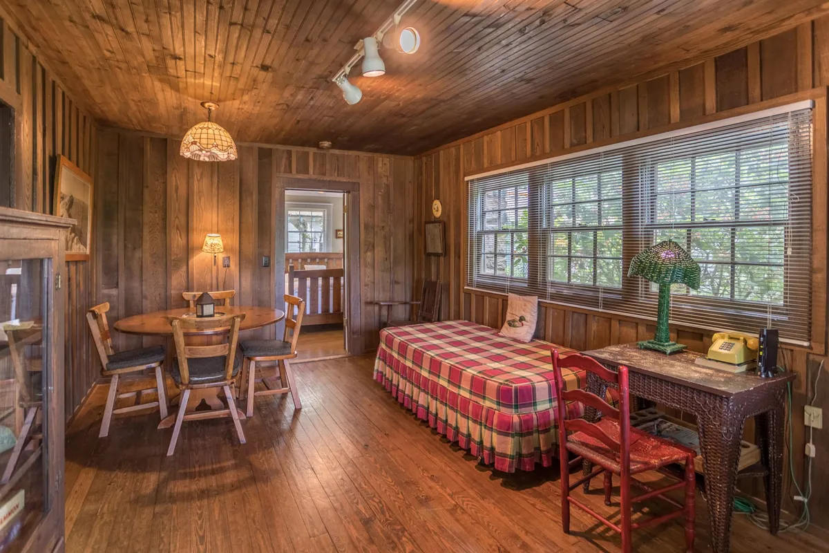 A cozy, wood-paneled room with a plaid bedspread on the bed, a dining table, and a woven lamp casting a soft glow. The window lets in morning light, and the scene radiates warmth and comfort, inviting guests to unwind and connect.