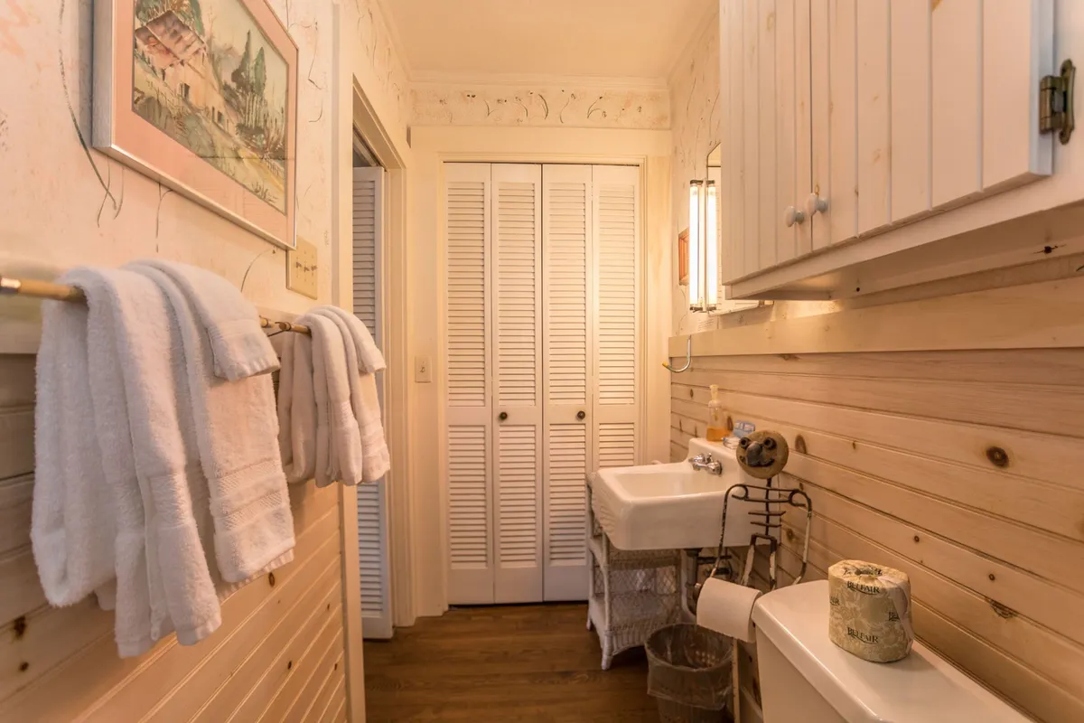 A thoughtfully designed bathroom with neatly hung towels, a sink with a vintage-style faucet, framed artwork on the wall, and a woven basket for storage. The atmosphere is tranquil and inviting, blending period charm with modern convenience.