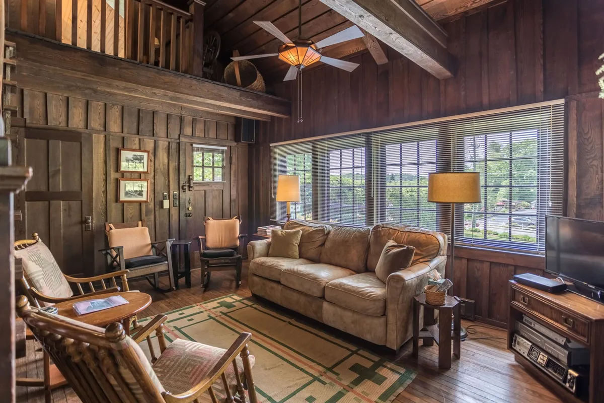 A cozy living area with a comfortable sofa, exposed wooden beams, large windows, and a decorative light fixture overhead. Rocking chairs positioned for relaxation and conversation, with soft natural light accentuating the rich wood details throughout the room.