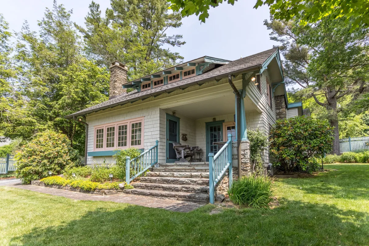 The image showcases the inviting exterior of a charming single-family cottage in Blowing Rock, North Carolina. Stone steps lead up through a beautifully landscaped garden to a front porch accented with colorful trim. The porch looks welcoming for relaxation, and the garden features a gazebo perfect for outdoor gatherings, all set against a lush backdrop.