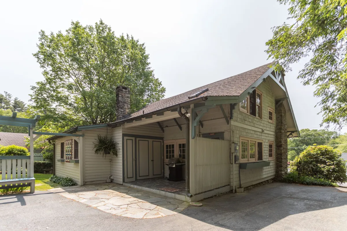 This exterior image showcases the home's stone chimney, broad eaves, large windows, and inviting front porch. The porch is furnished and set against a lush garden backdrop, hinting at both curb appeal and the comfort of outdoor living spaces. The surrounding greenery and yard are visible, evoking the serenity and privacy described in the section.