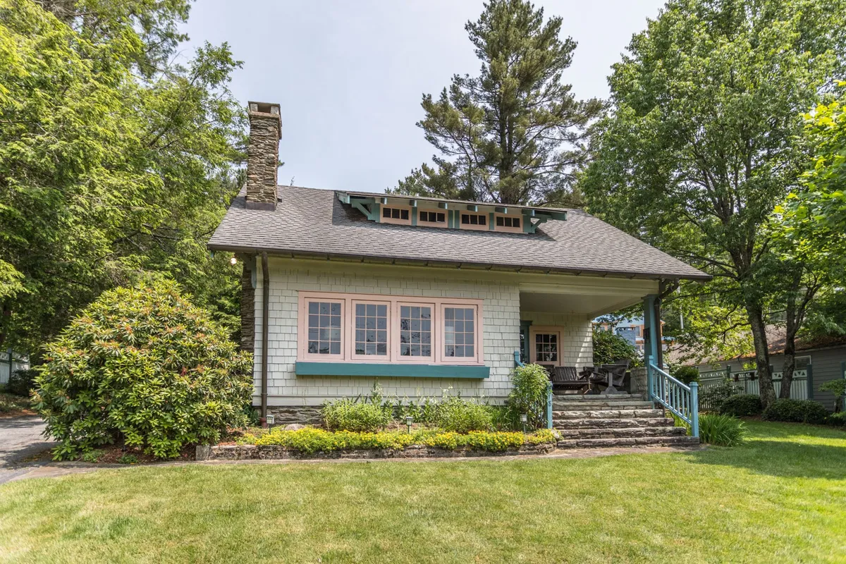 This image presents the broad front porch with its stone chimney and large windows, framed by greenery. The perspective offers a sense of both openness and privacy, with seating for relaxation and ample space for gatherings or morning coffees. The porch's connection to the landscape is evident, blending indoor comfort with outdoor enjoyment.