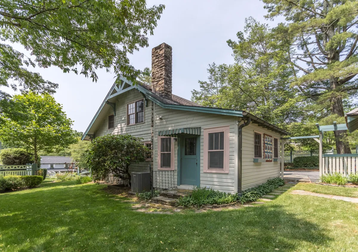 This image features the charming exterior of the Suddreth Cottage, with its inviting stone chimney, colorful window trim, and lush green lawn. The scene radiates warmth and historic character, capturing the welcoming essence of a well-kept, early 1900s bungalow surrounded by greenery. The exterior hints at the home's historic bones and the experience of stepping into a storied past.