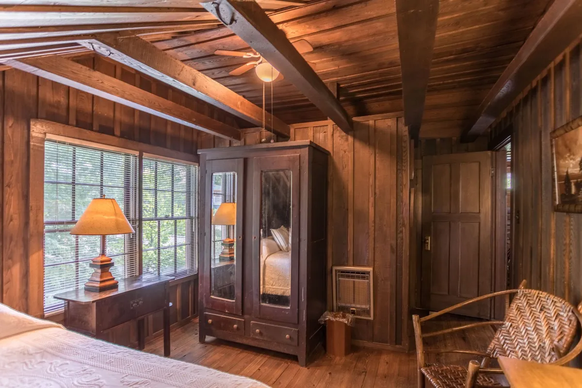 A nostalgic bedroom featuring exposed wooden beams, an antique wardrobe, a soft bedside lamp, and a woven chair. The bed is set in a tranquil corner with vintage décor and natural morning light creating a peaceful, inviting space.