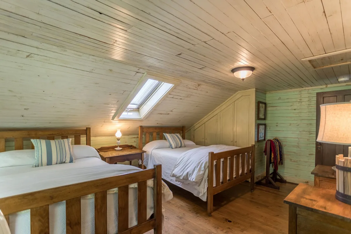An inviting upstairs bedroom with two wooden beds positioned beneath a skylight, a central nightstand with a lamp, and gentle natural light illuminating the room. The setting is cozy and relaxed, encouraging restful sleep and quiet moments for families or friends.