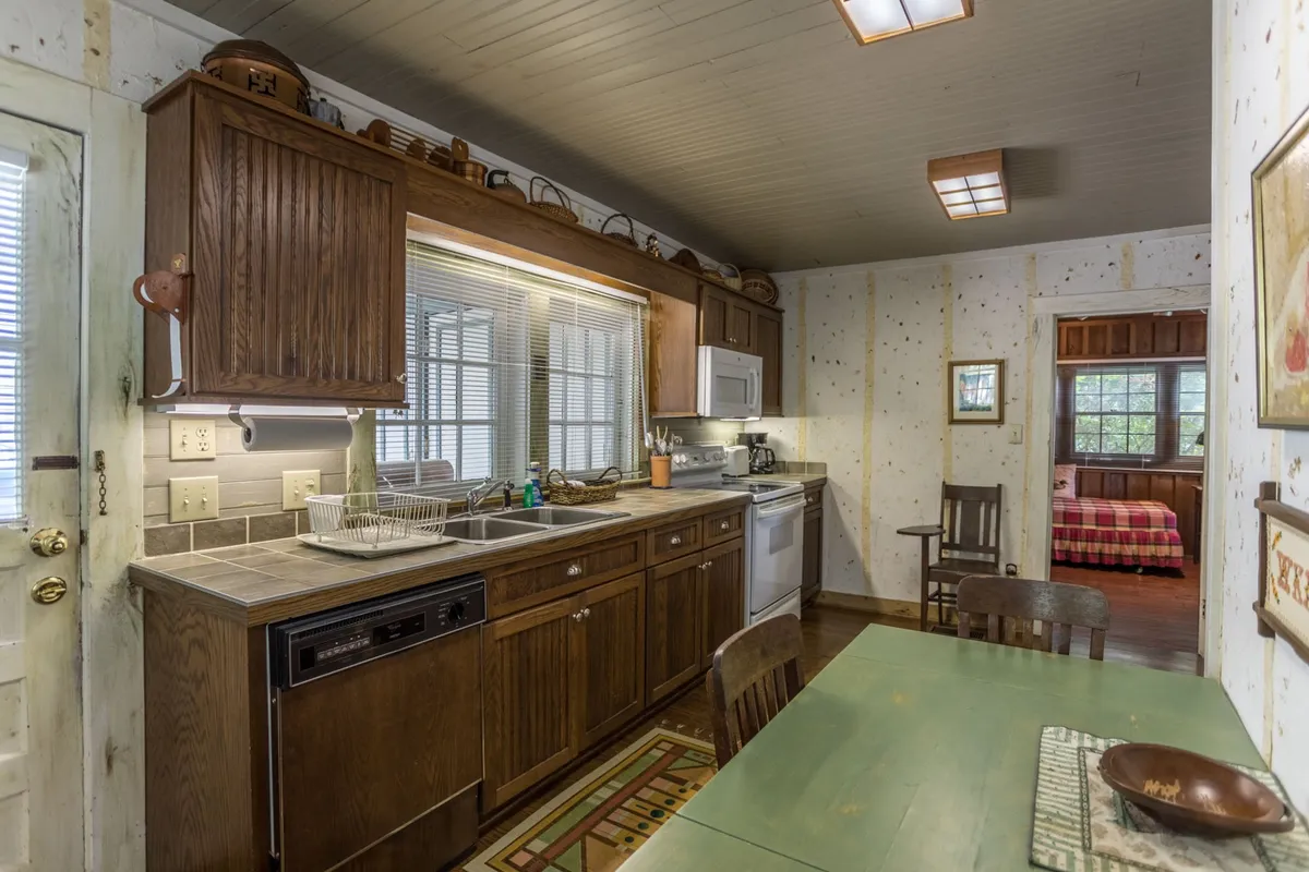 This image centers on a warm, homey kitchen with wooden cabinetry, a kitchen island, a dining table, and sunlit windows. The space is set for cooking, gathering, and sharing meals, with subtle artistic details visible in the decor.